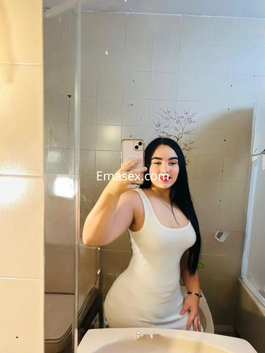 Puta en Sevilla posando frente al espejo con un vestido blanco ajustado y cabello largo y negro.