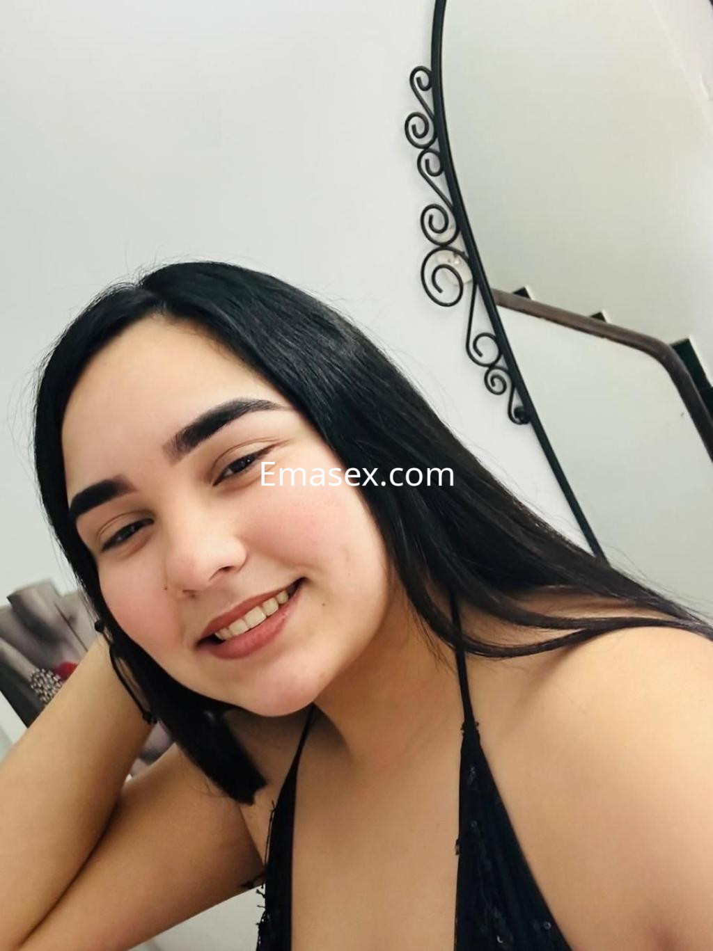 Escort en Sevilla con cabello largo y oscuro, sonriente y posando en un entorno interior con espejo.