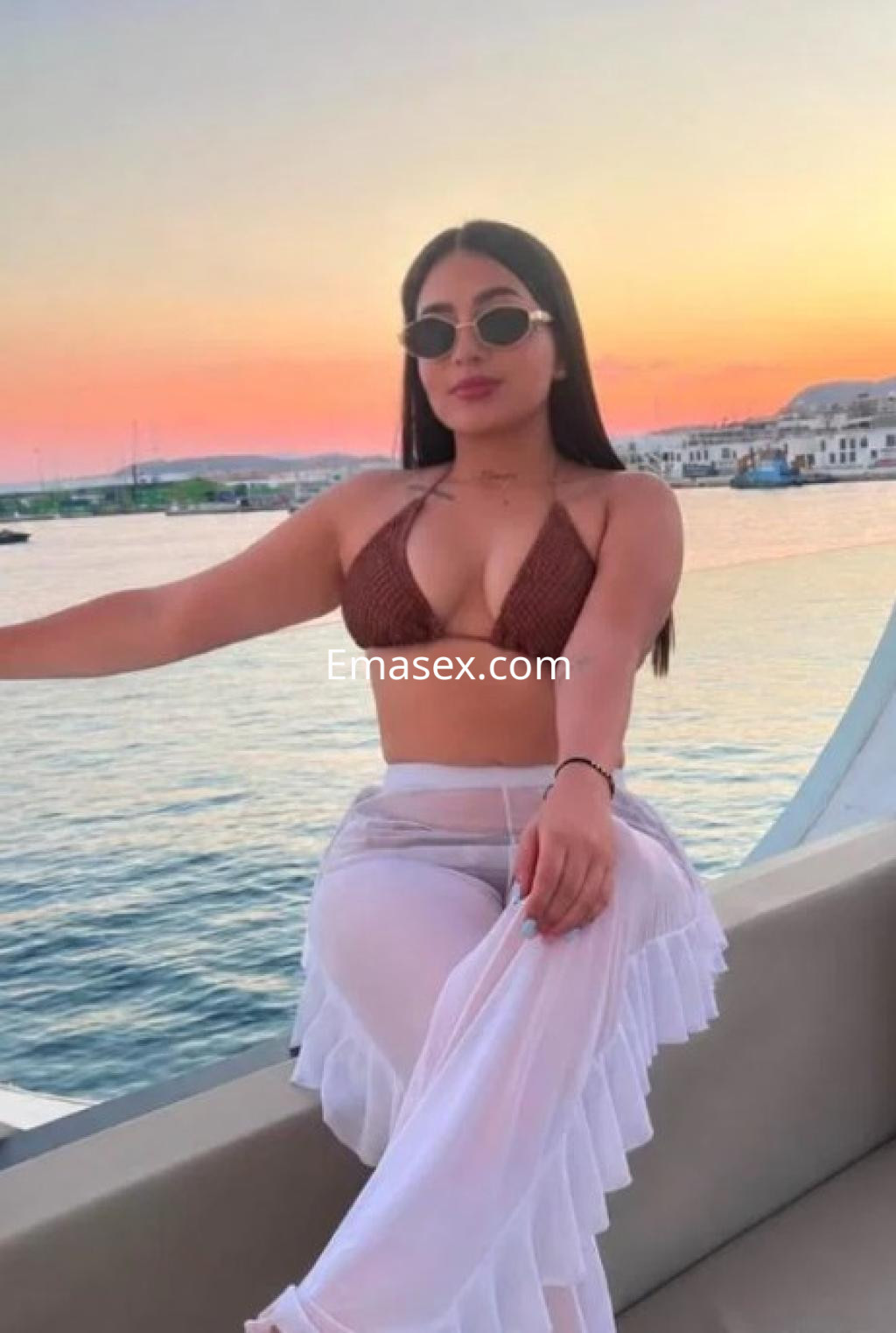 Escort en Castellón, posando en un yate con un bikini marrón y una falda blanca, al atardecer junto al mar.