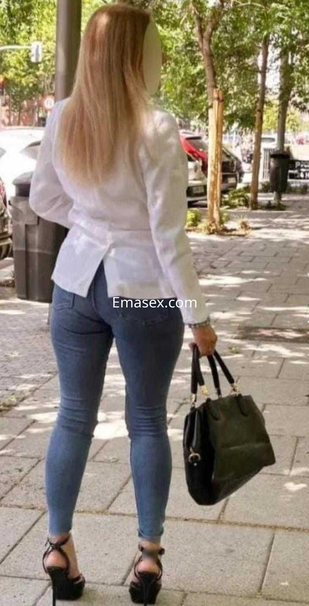 Escort en Castellón, con cabello rubio y lacio, viste blusa blanca y jeans ajustados, caminando por la acera.