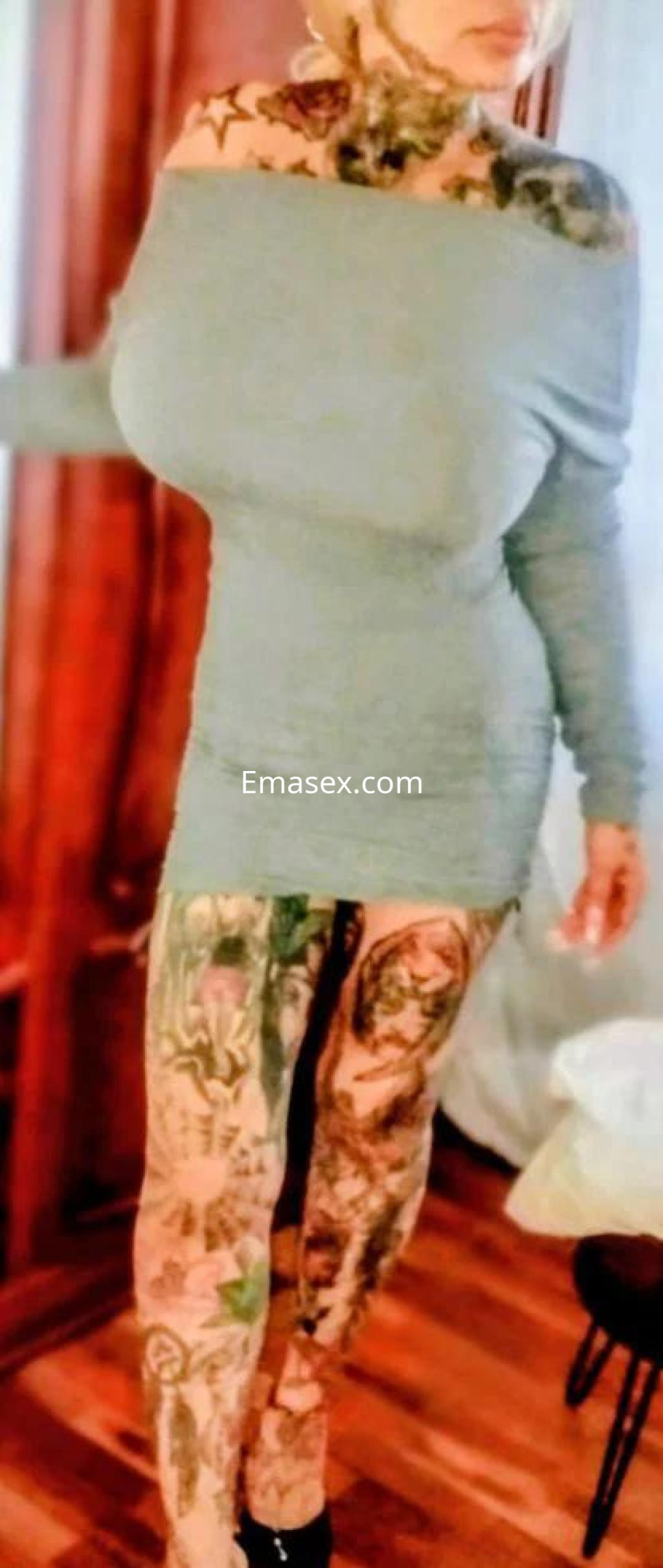 Escort de Ourense de pie, viste un vestido ajustado de color gris claro y tiene tatuajes en brazos y piernas.