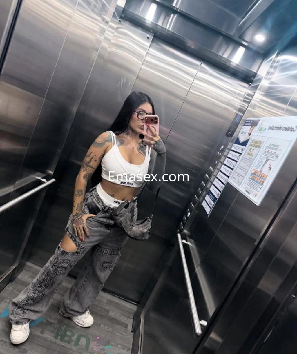 Escort en Ourense posando en un ascensor, viste un top blanco y pantalones grises rasgados, con tatuajes visibles.