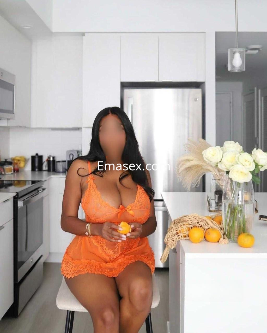 Escort en Cáceres, sentada en una cocina moderna, viste lencería naranja y sostiene una mandarina.