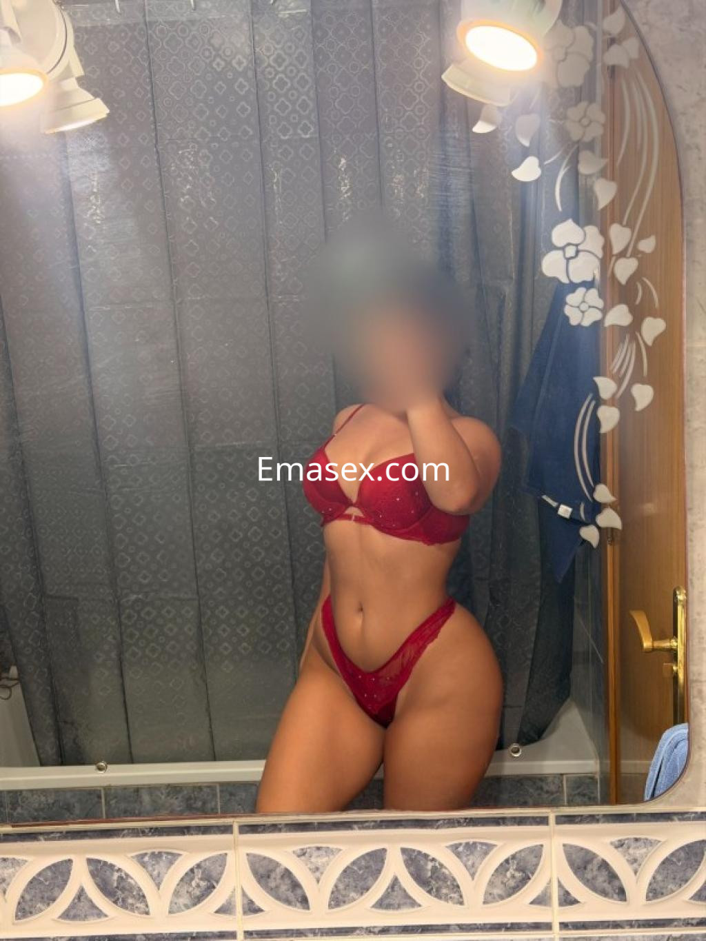 Puta en Tarragona posando con lencería roja frente a un espejo en un baño, mostrando su figura curvilínea.