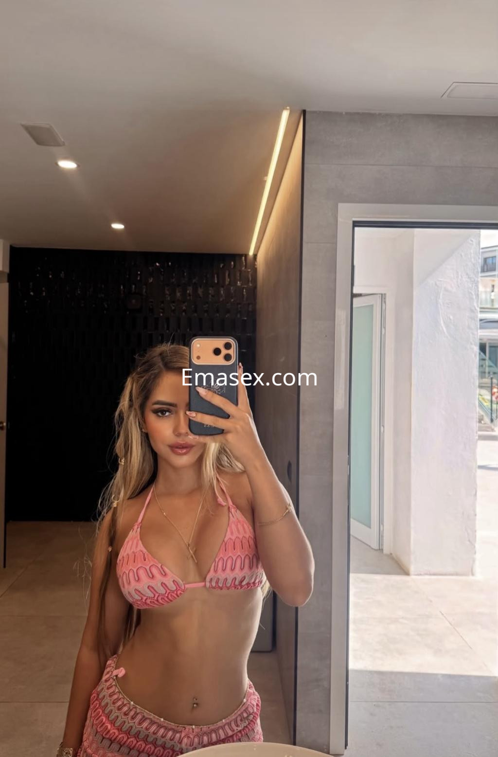 Puta en Vigo, con cabello largo y suelto, luciendo un bikini rosa y posando frente a un espejo moderno.