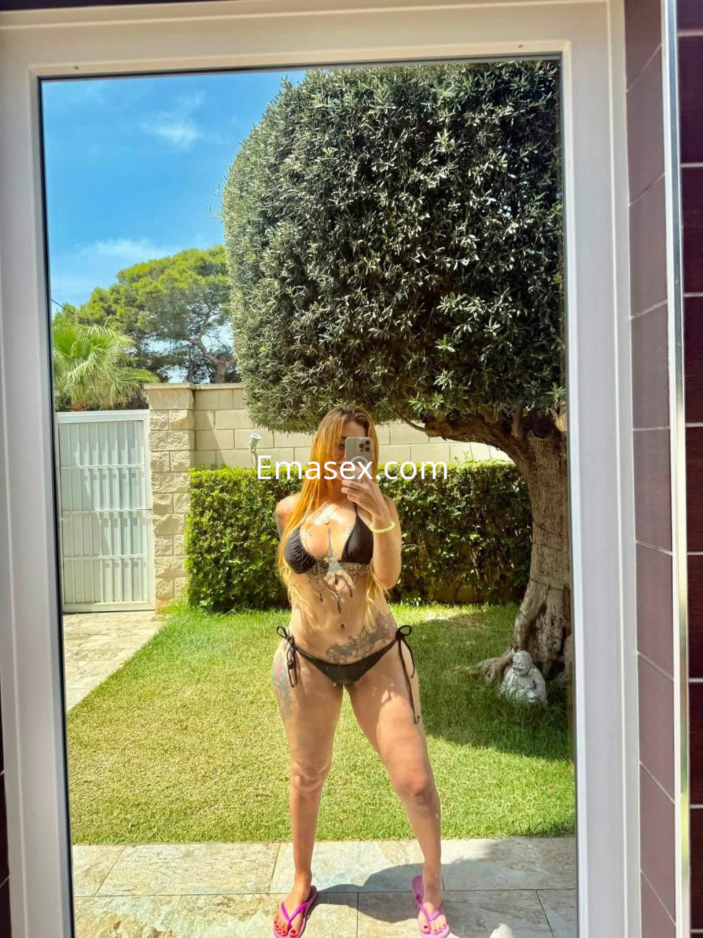 Escort de cabello castaño largo, con un bikini negro, posando frente a un espejo en un jardín en Pamplona.