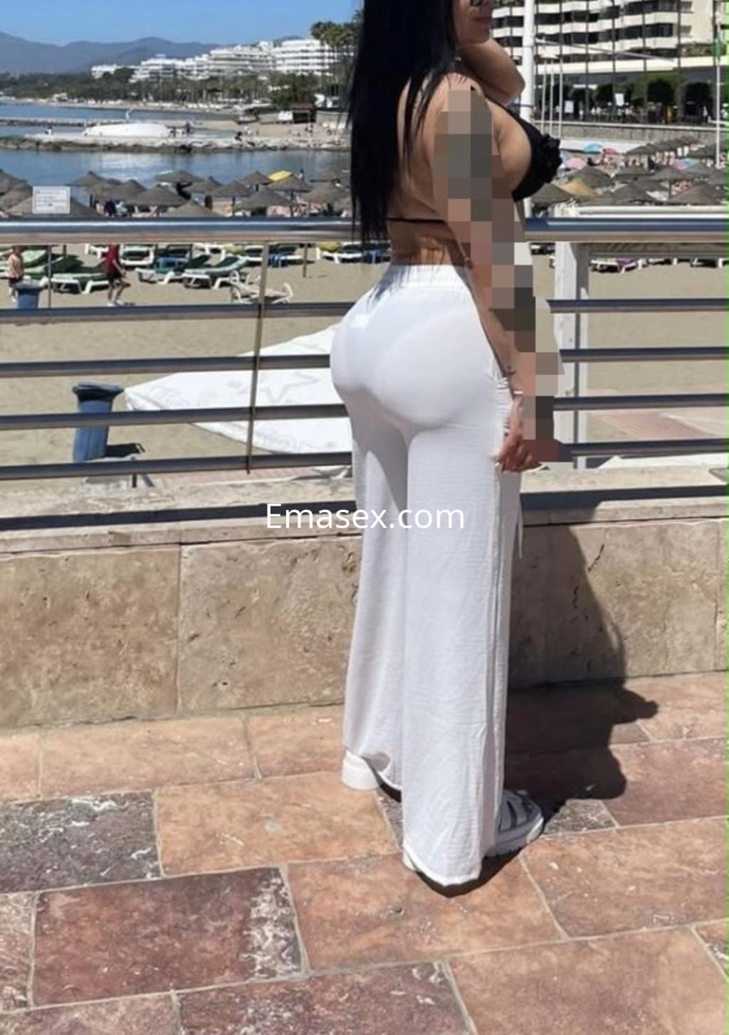 Puta en Pamplona con cabello largo y oscuro, usando top negro y pantalones blancos, posando en la playa.