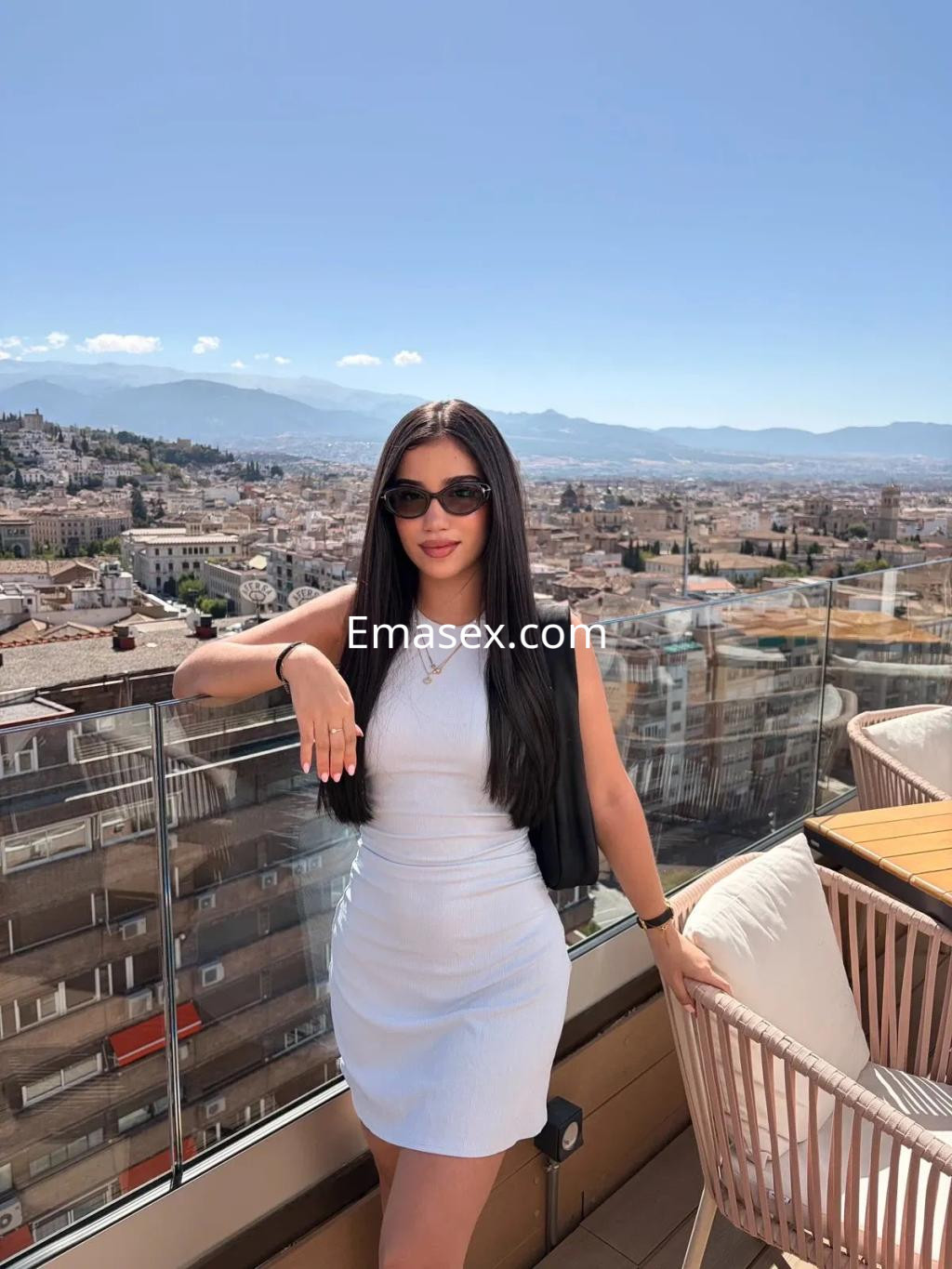 Escort en Zaragoza con vestido blanco ajustado y chaqueta negra, posando con gafas de sol en una terraza con vista.