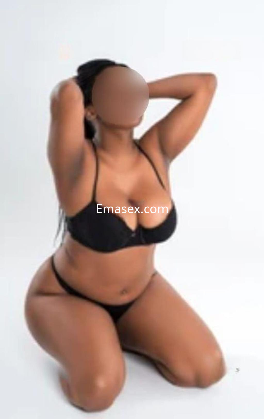 Escort posando de rodillas en lencería negra, con cabello oscuro y piel morena, en un fondo blanco en Pontevedra.