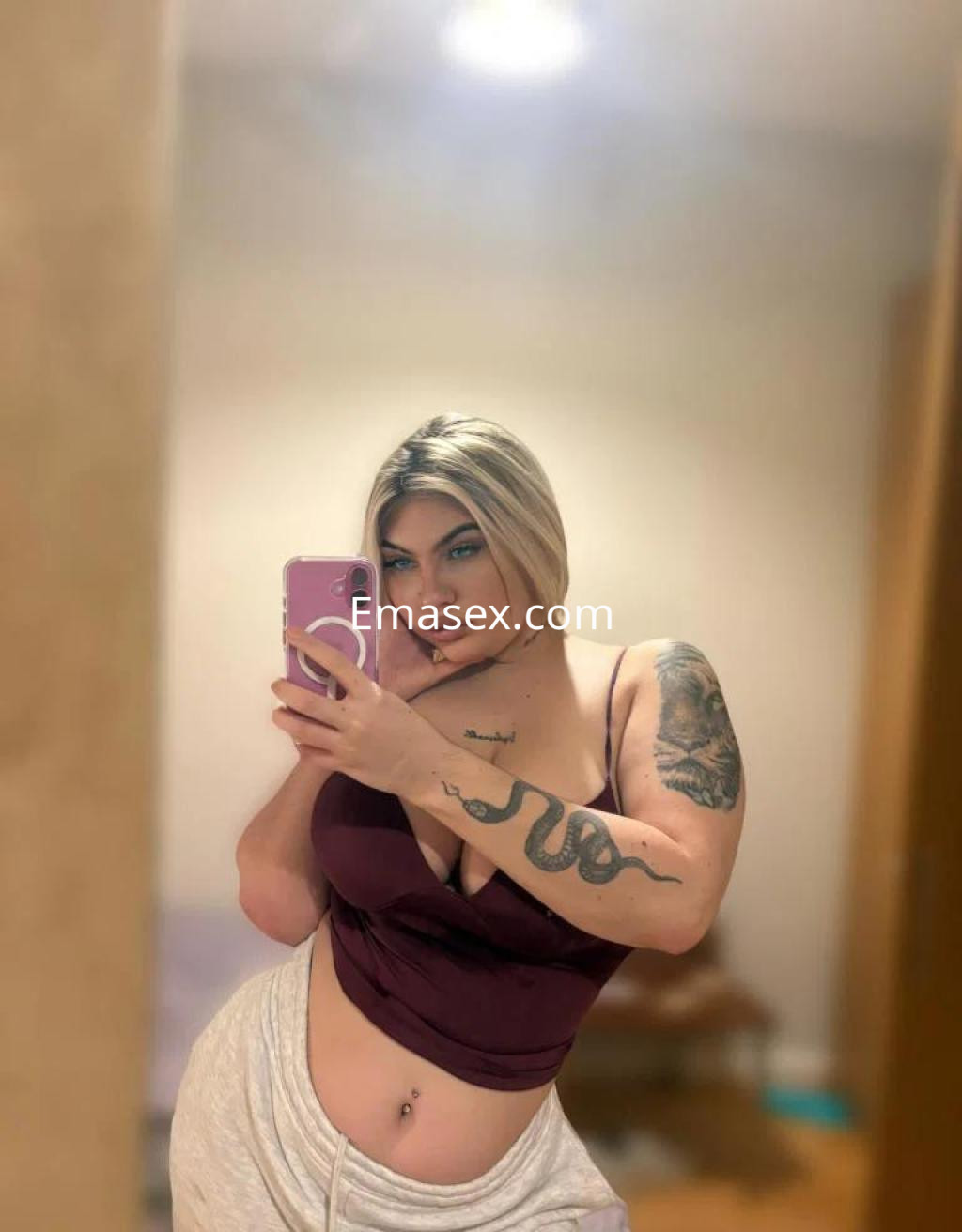 Escort en Coruña posando frente al espejo, con blusa sin mangas y pantalones claros, mostrando tatuajes.