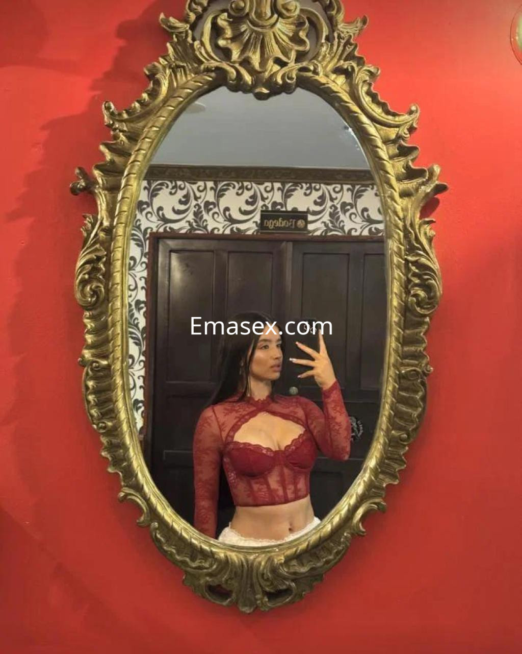 Puta en Coruña posa frente a un espejo con un top de encaje rojo y pantalones claros en un ambiente decorativo.