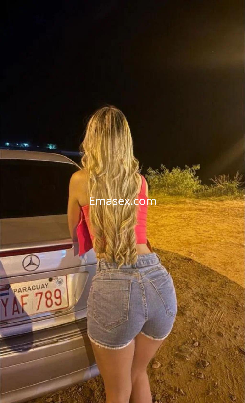 Escort con cabello largo y rubio, luciendo un top rosa y shorts de mezclilla, apoyada en un coche en Castellón.