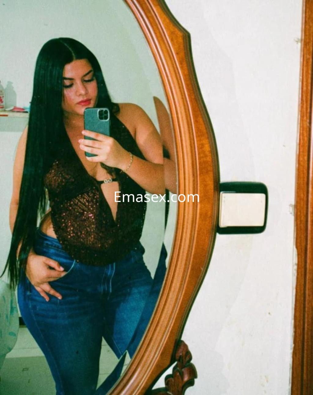 La escort en Badajoz se toma una selfie frente a un espejo, luciendo una blusa brillante y jeans ajustados.