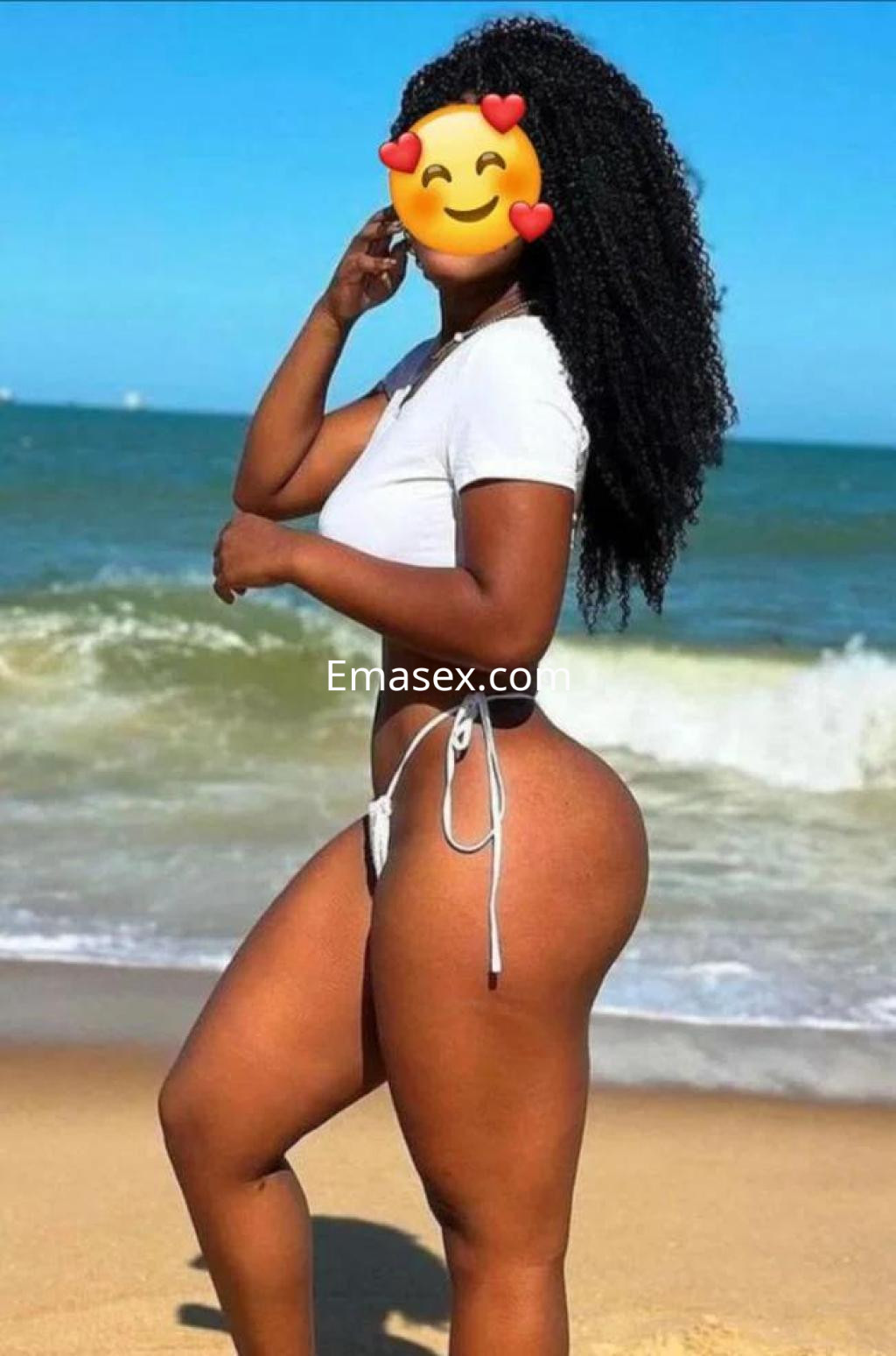 Escort en Murcia con cabello rizado, luce un bikini blanco mientras posa frente al mar en la playa.