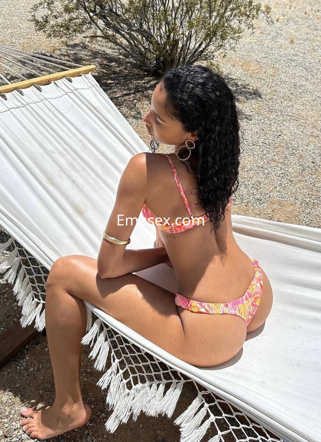 Puta en Ibiza, posando de espaldas en una hamaca con un bikini de colores vivos y cabello rizado.