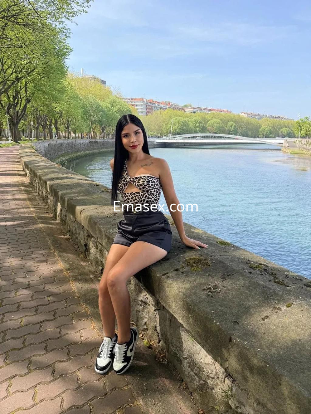 Puta sentada sobre un muro junto al río en Coruña, vistiendo un top de leopardo y pantalones cortos oscuros.