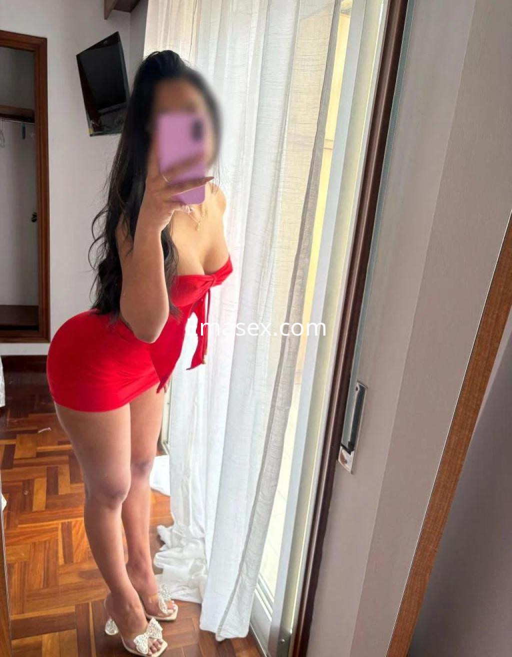 La puta luce un vestido rojo ajustado y tacones, posando frente a una ventana en Ourense.