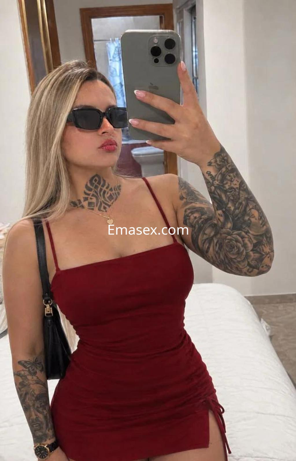 Escort en Las Palmas con vestido rojo ajustado, gafas de sol y tatuajes visibles en los brazos, posando frente al espejo.