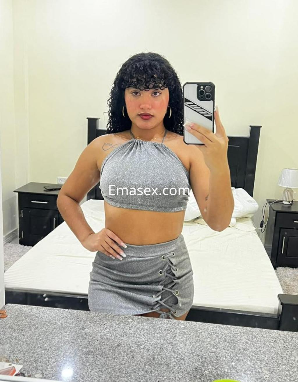 Puta en Las Palmas, con cabello rizado y oscuro, viste un top plateado y una falda corta, posando frente a un espejo.