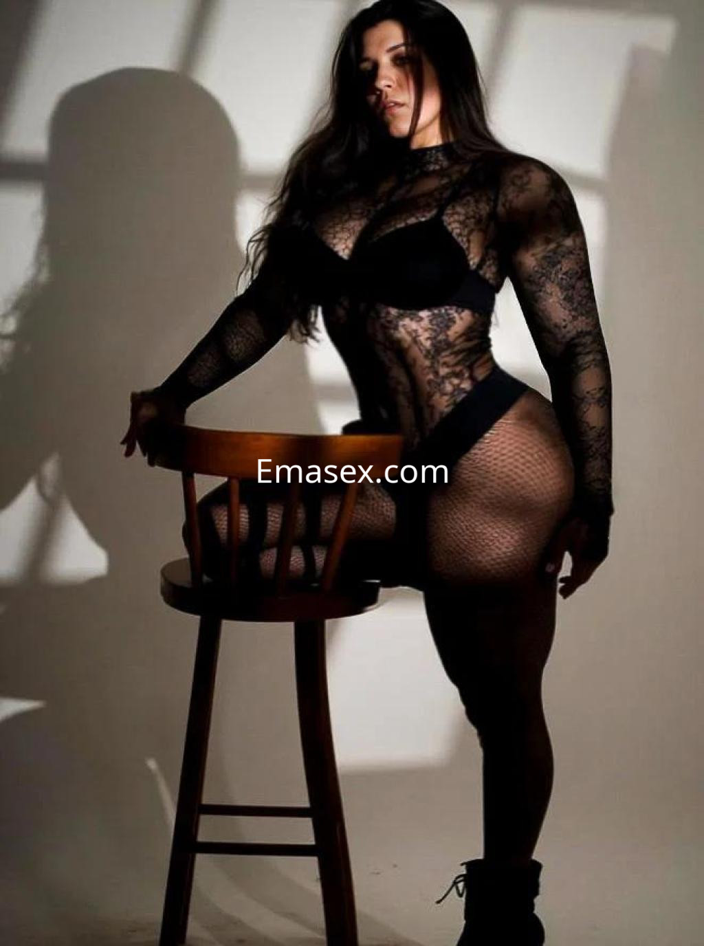 Puta en Lugo posando con una malla negra de encaje y medias de red, apoyada en una silla de madera.