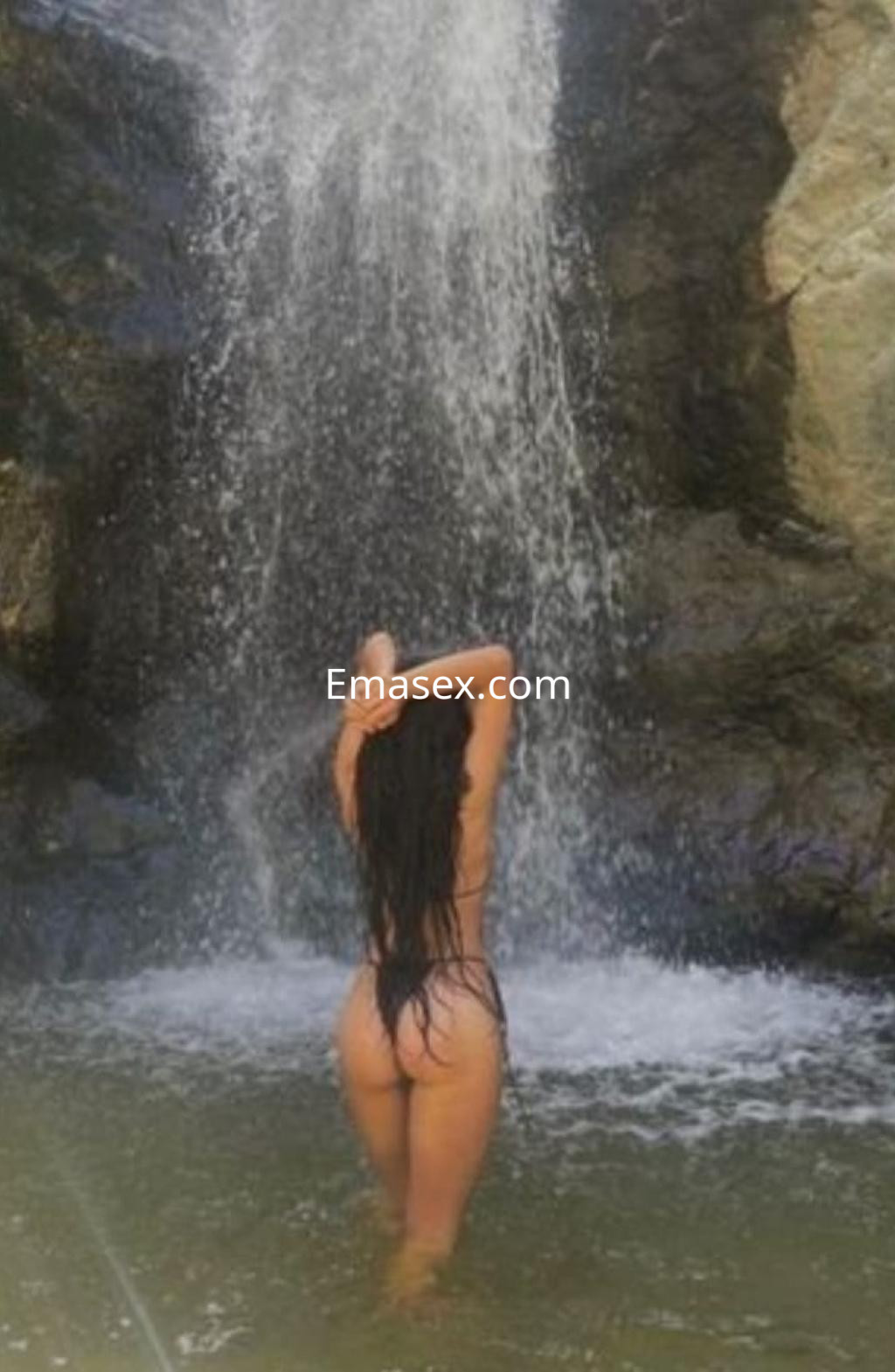 La escort en Zaragoza se encuentra de espaldas en una cascada, luciendo un bañador negro y mojándose el cabello.