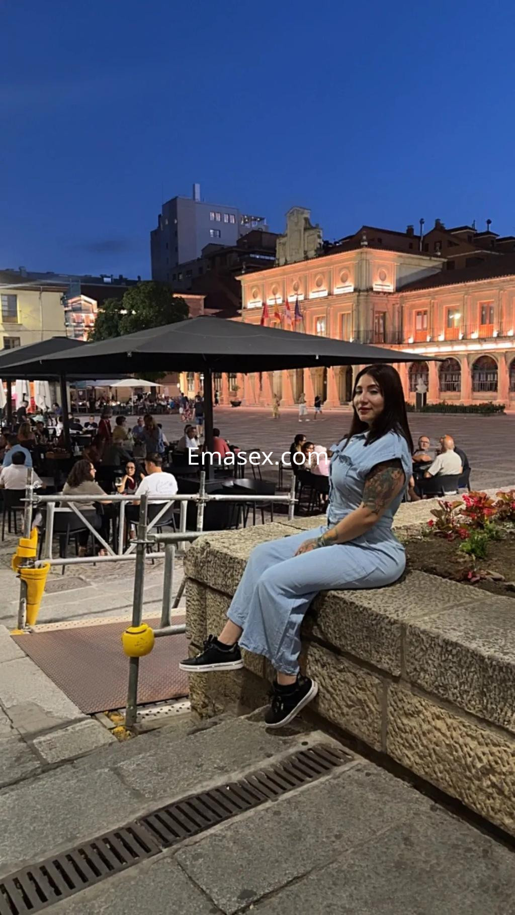 Escort sentada en un muro, luciendo un overol de denim y zapatillas, con un entorno urbano de Zaragoza al anochecer.