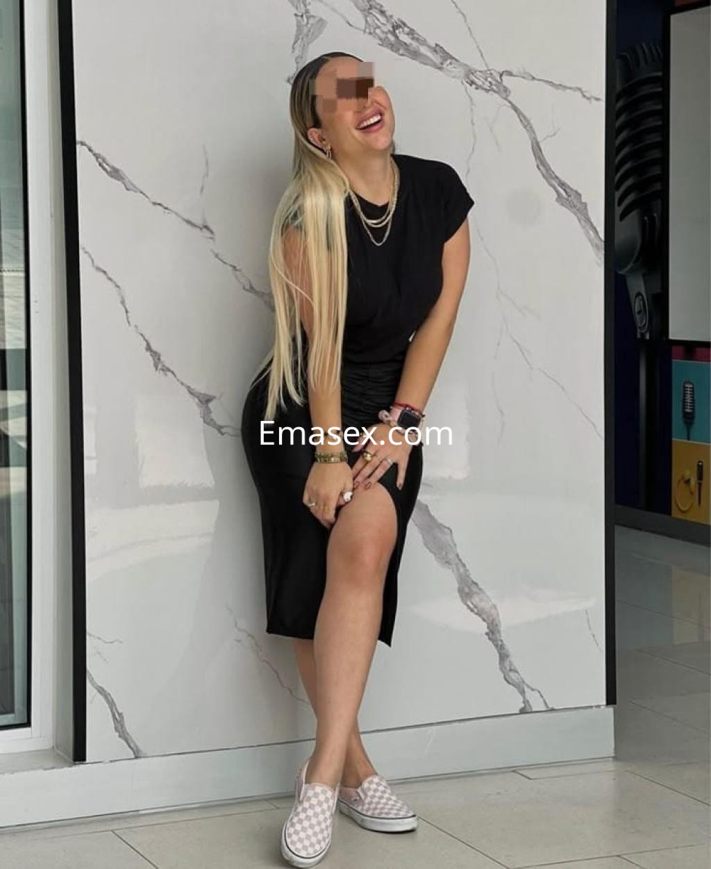 Escort en Bilbao, con cabello largo y lacio, viste un vestido negro y zapatos de lona, sonriendo frente a una pared.