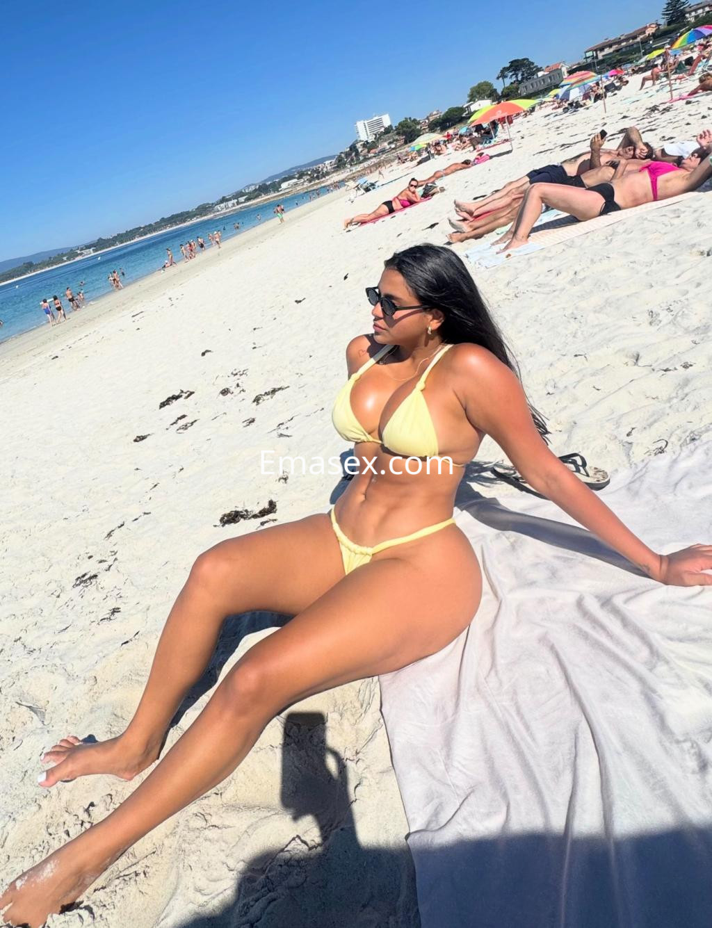 Puta sentada en la playa de Vigo, luciendo un bikini amarillo y gafas de sol, con el mar de fondo.