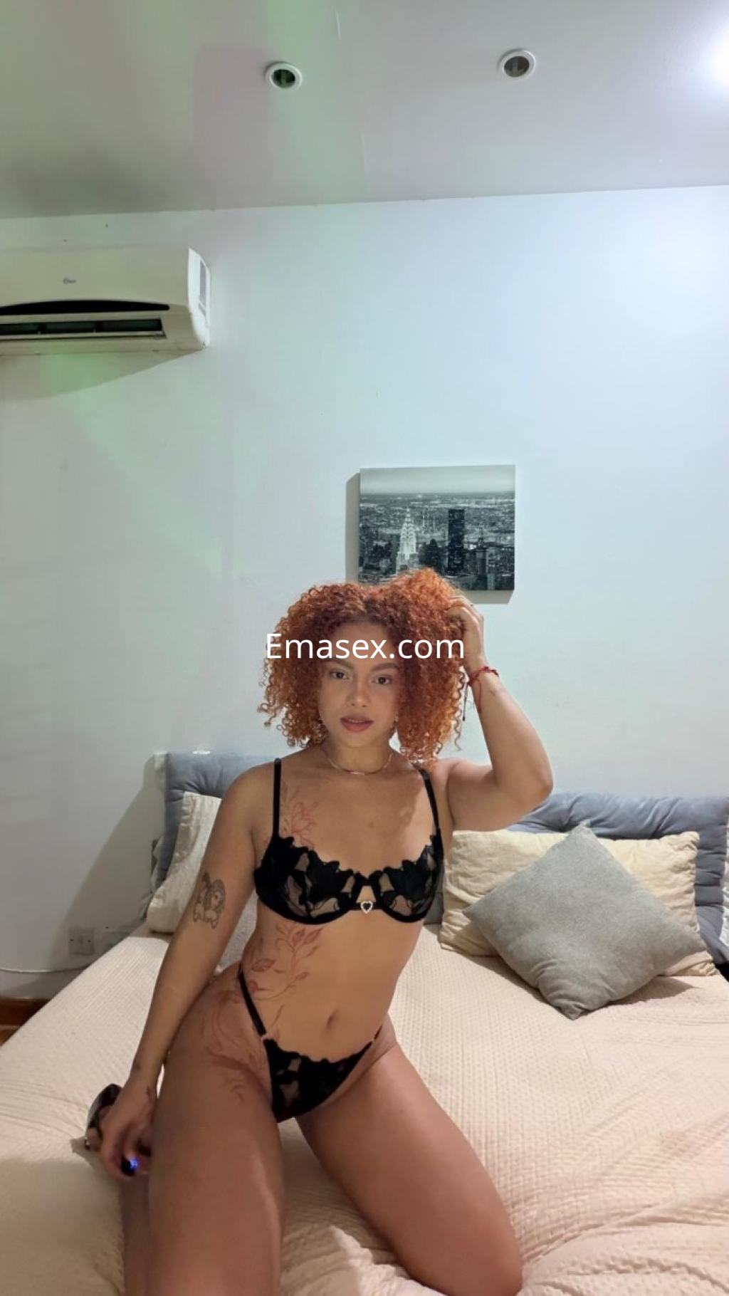 Escort en Girona, con cabello rizado y pelirrojo, posa en lencería negra sobre una cama en un ambiente acogedor.