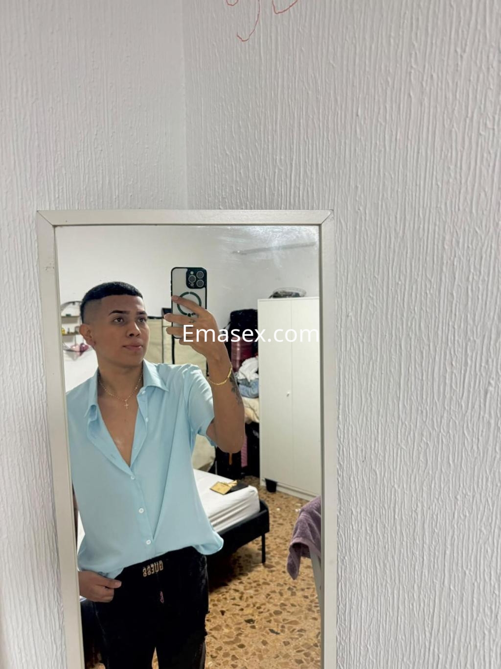 Puta trans en Bilbao frente al espejo, luciendo una camisa celeste y pantalones oscuros en un entorno doméstico.