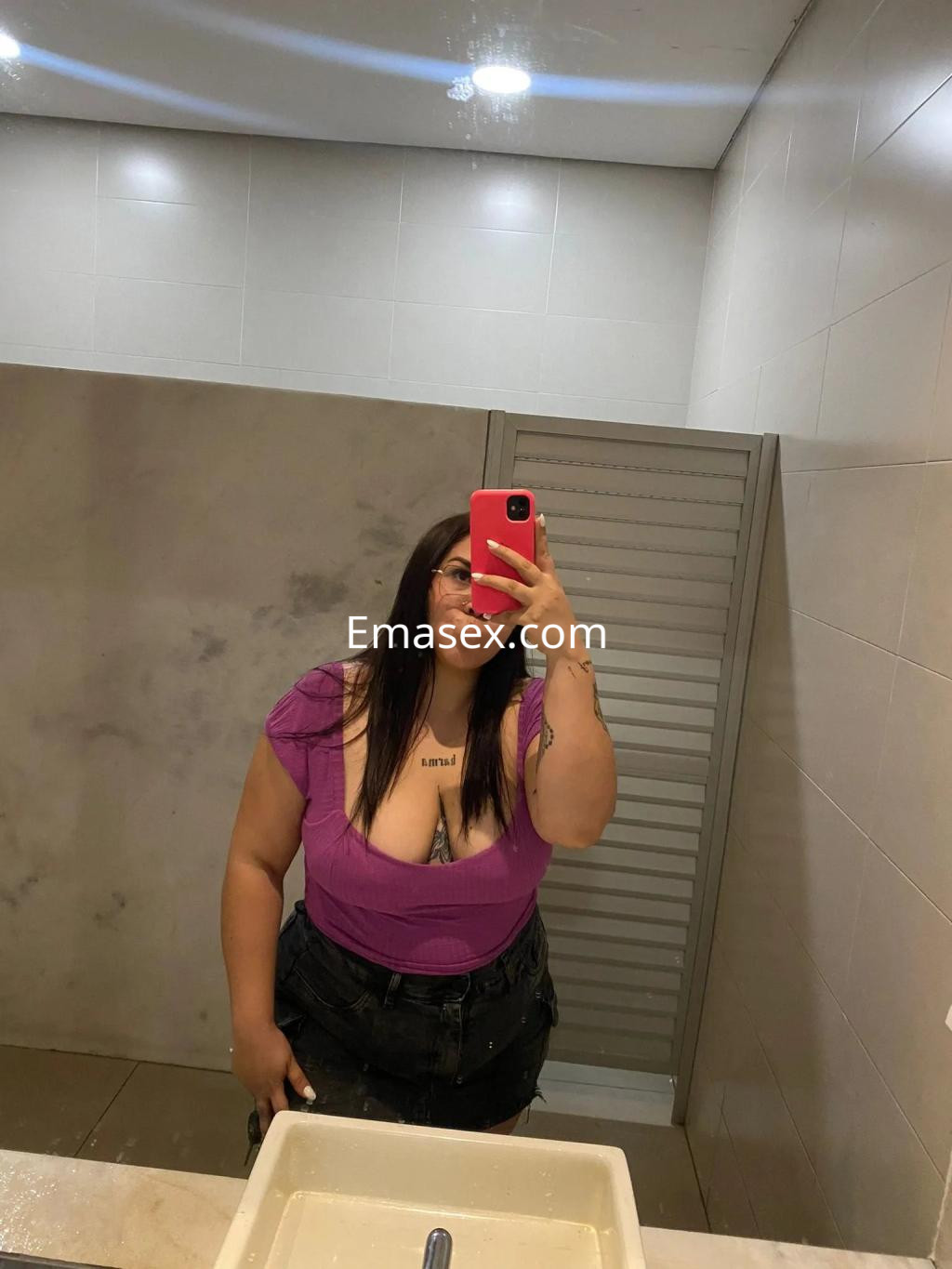 Escort en Zaragoza con cabello largo y castaño, luciendo una blusa morada y shorts de mezclilla en un baño.