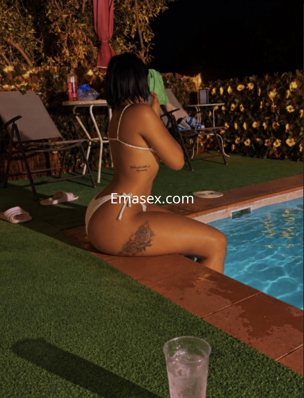 Escort en Donosti sentada al borde de una piscina, luciendo un bikini blanco y mostrando un tatuaje en la pierna.
