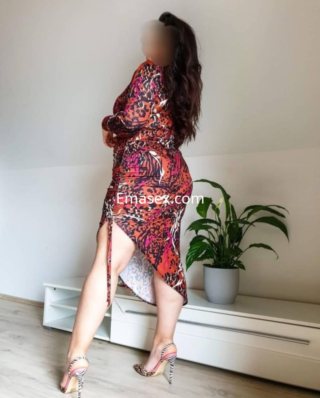 Escort posando de espaldas en Girona, con vestido colorido y zapatos de tacón, en un interior moderno.