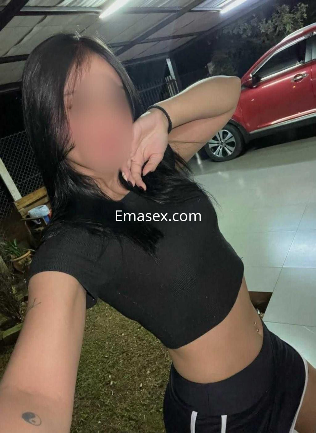 Escort en Ibiza con cabello negro, lleva una camiseta corta y una falda negra, posando en un entorno exterior nocturno.