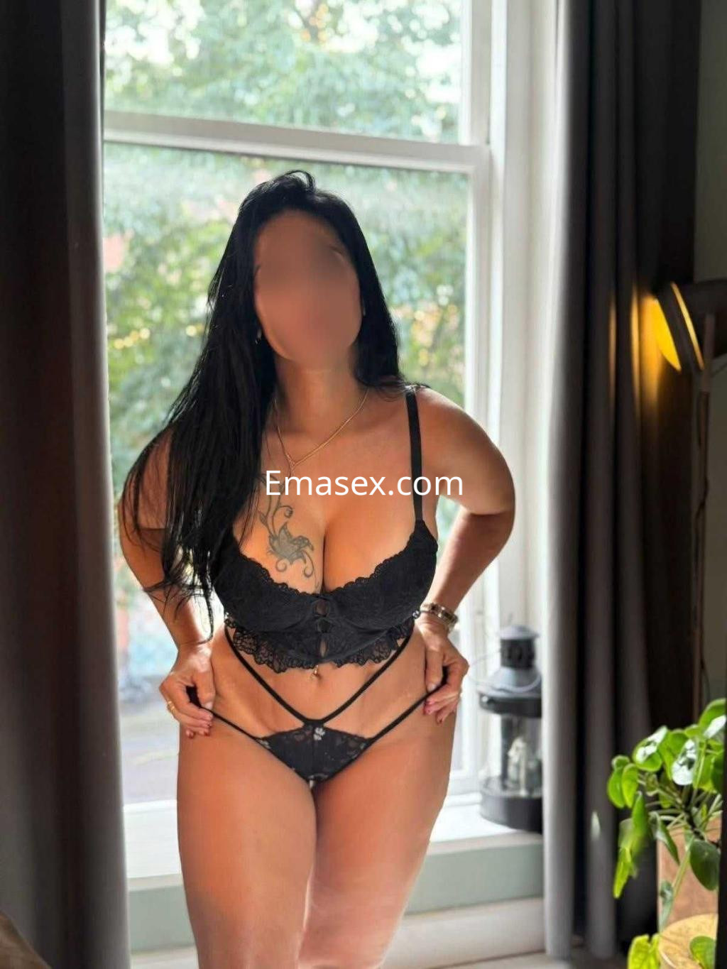 Puta con lencería negra ajustada, posando en una habitación iluminada con ventana, en Ibiza.