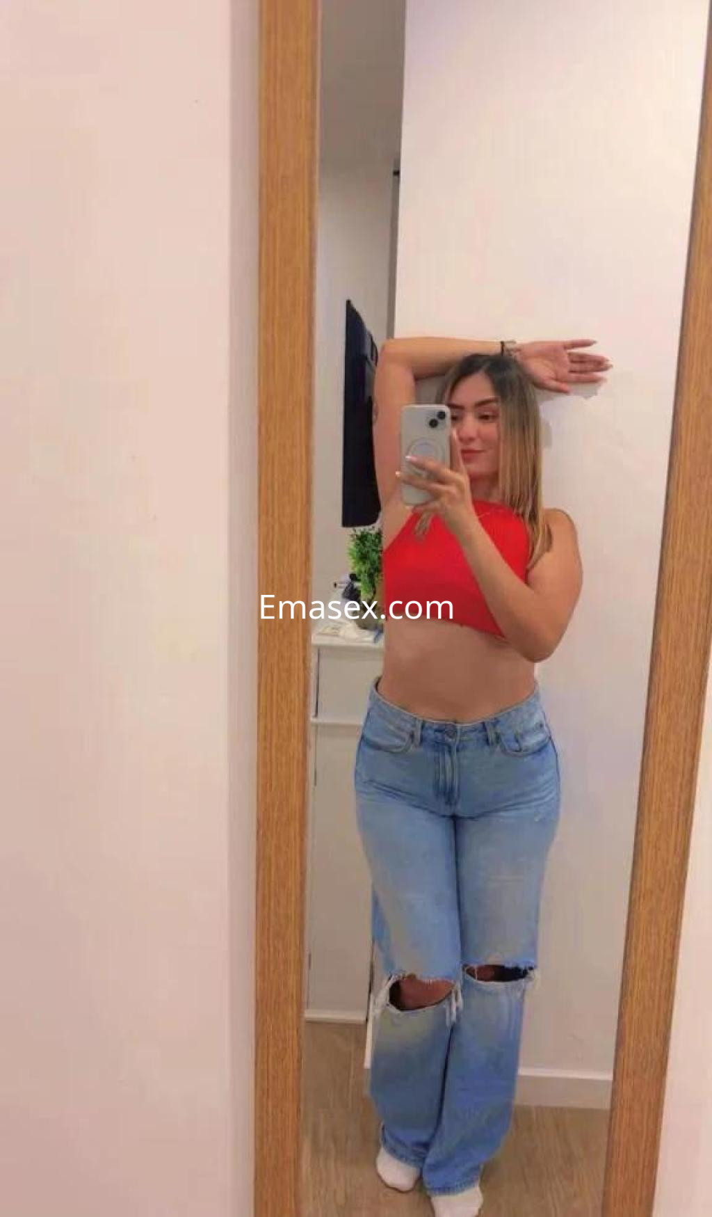 Escort en Pontevedra posa en un espejo, vistiendo un top rojo y jeans rasgados, con cabello largo y lacio.