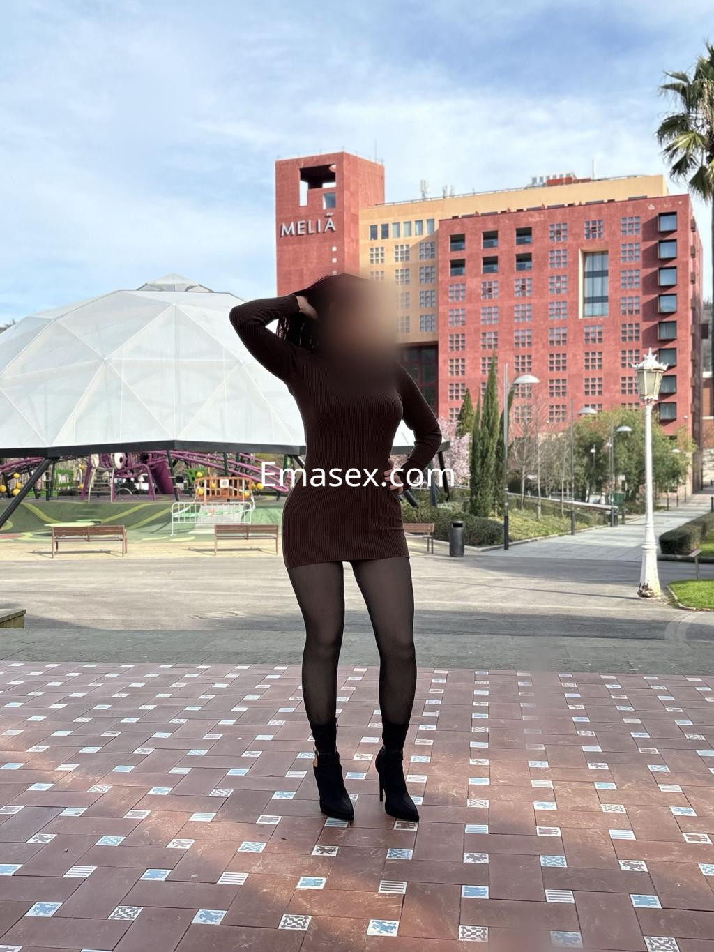 Escort en Bilbao, con un vestido ajustado marrón y botas negras, posando en un parque con edificios al fondo.