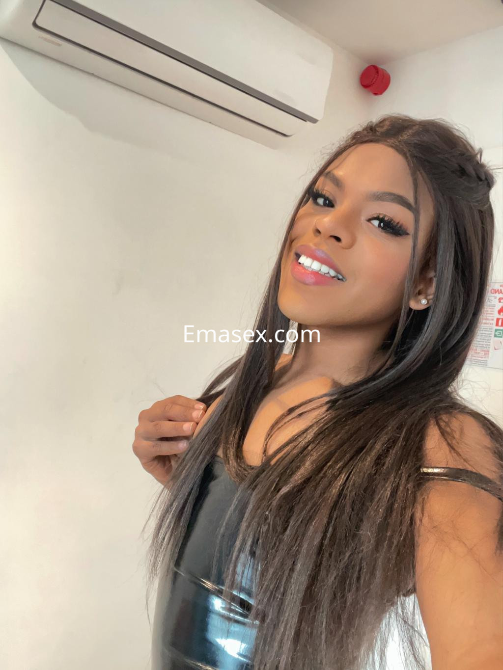 Escort trans en Irún con cabello largo y lacio, vistiendo un vestido negro brillante, sonríe en un entorno luminoso.