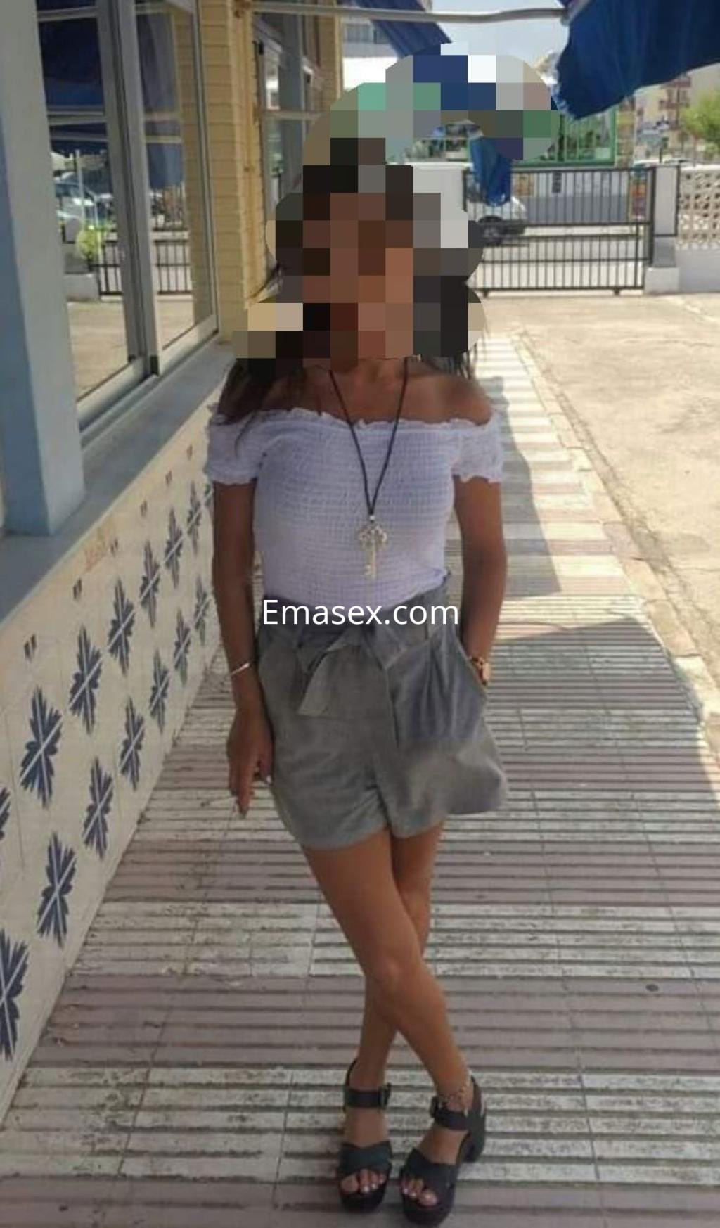 Escort en Valencia viste blusa blanca de hombros descubiertos y shorts grises, con sandalias negras y collar.