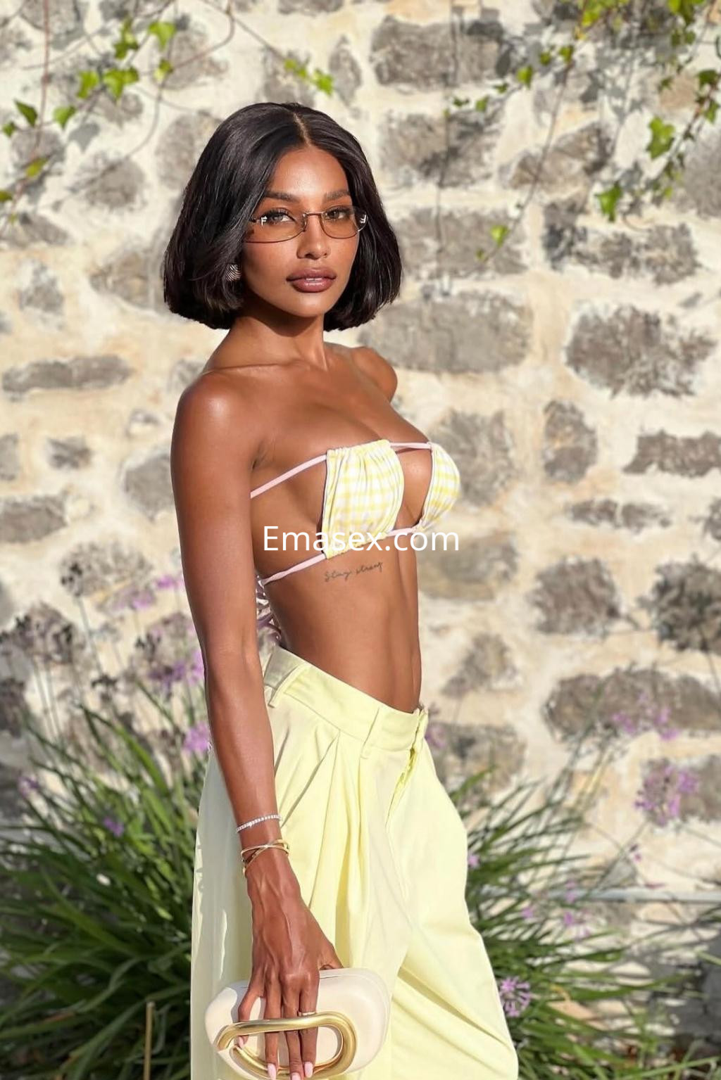 Escort en Ibiza con top de cuadros amarillos y pantalones amarillos, posando con gafas y un bolso claro.