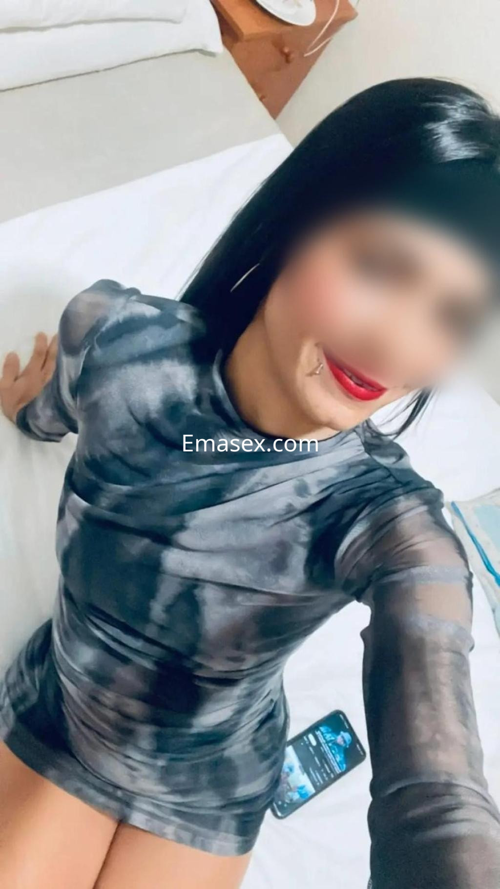 Escort en Pamplona, posando sentada en una cama con un vestido de malla negro y labios rojos.