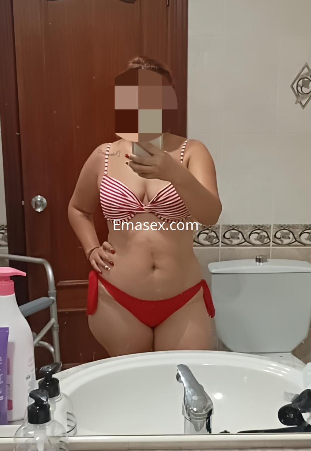 La puta en Lleida posa frente al espejo con un bikini de rayas rojas y blancas, mostrando su figura.