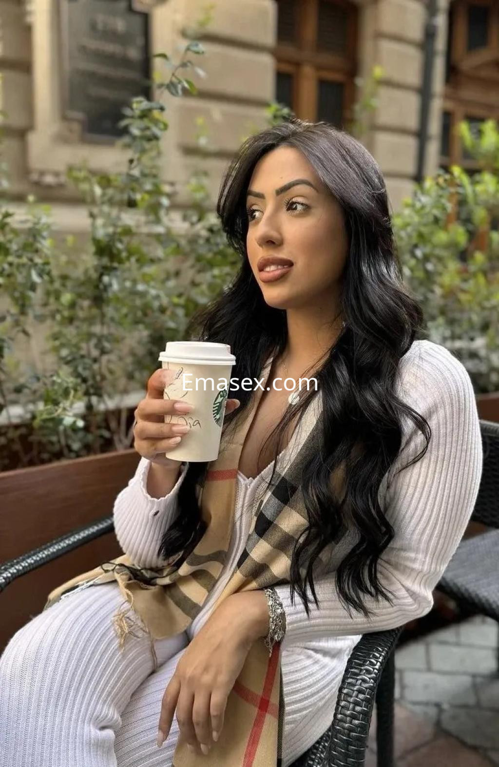 La puta lleva un abrigo blanco y una bufanda de cuadros mientras sostiene un café en una terraza de Alicante.