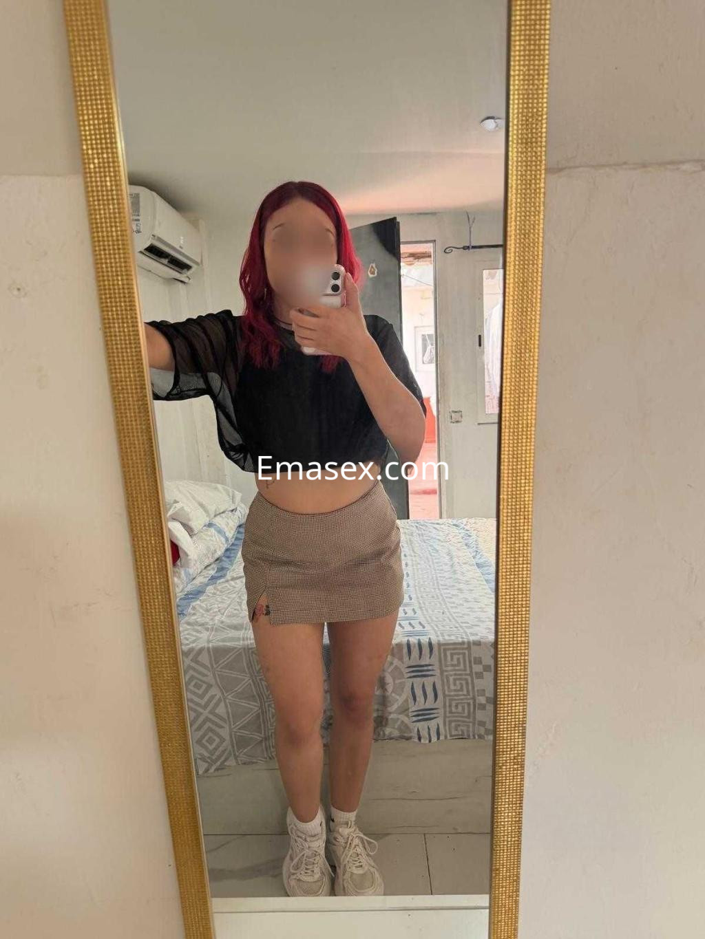 La escort en Castellón se muestra frente al espejo con una blusa negra y una falda corta de cuadros.