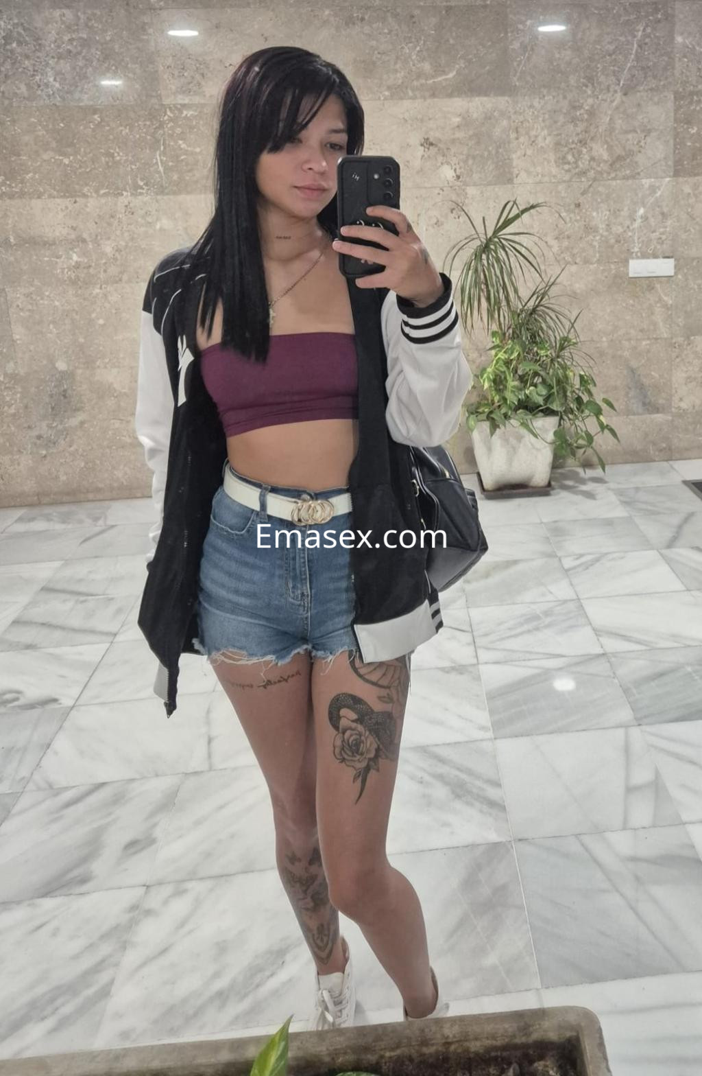 Escort en Sevilla con top burdeos y chaqueta negra, luciendo pantalones cortos de mezclilla y accesorios en un entorno moderno.
