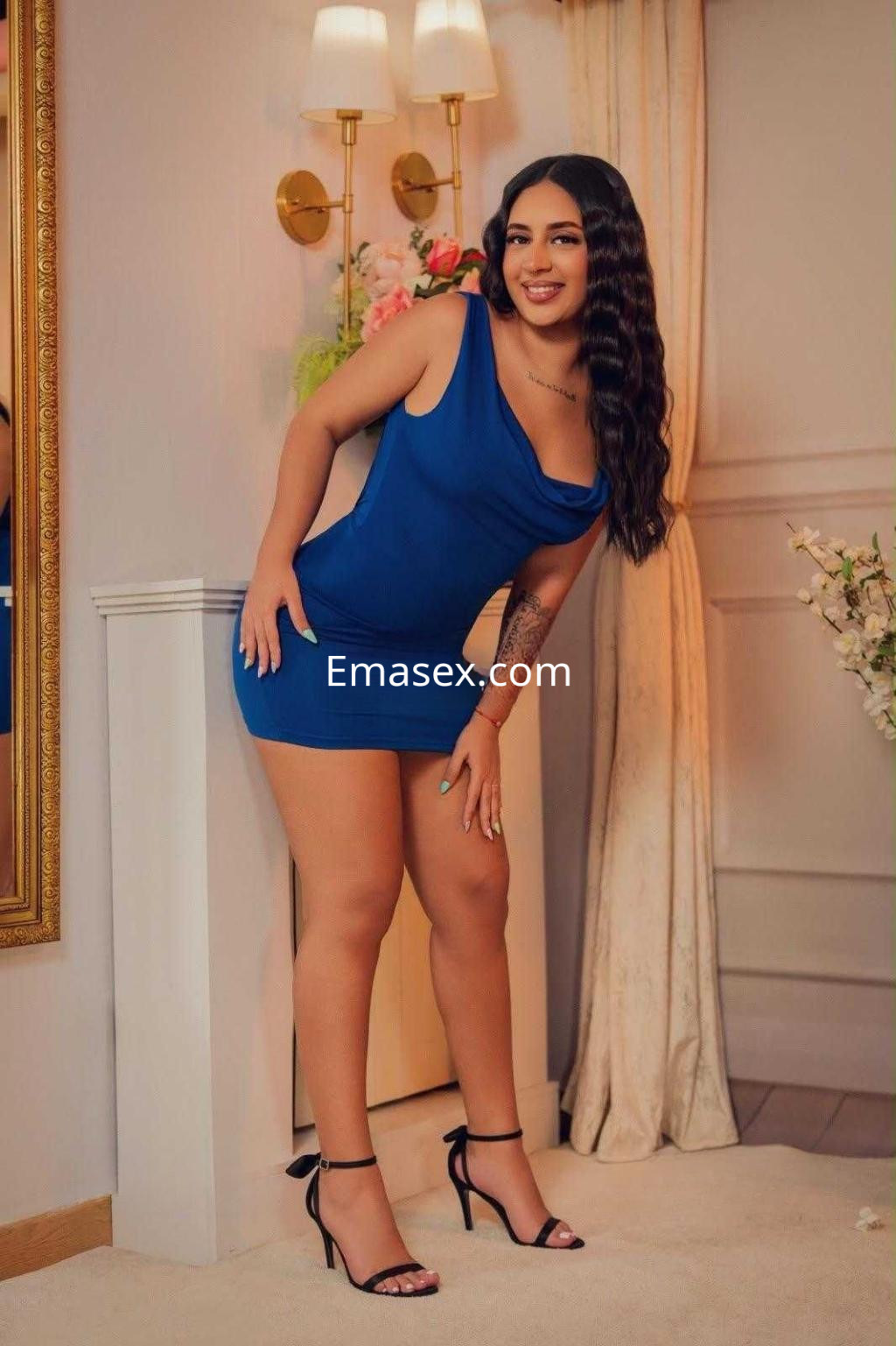 Escort en Ibiza posando con un vestido azul ajustado y tacones negros, sonriendo en un ambiente elegante.
