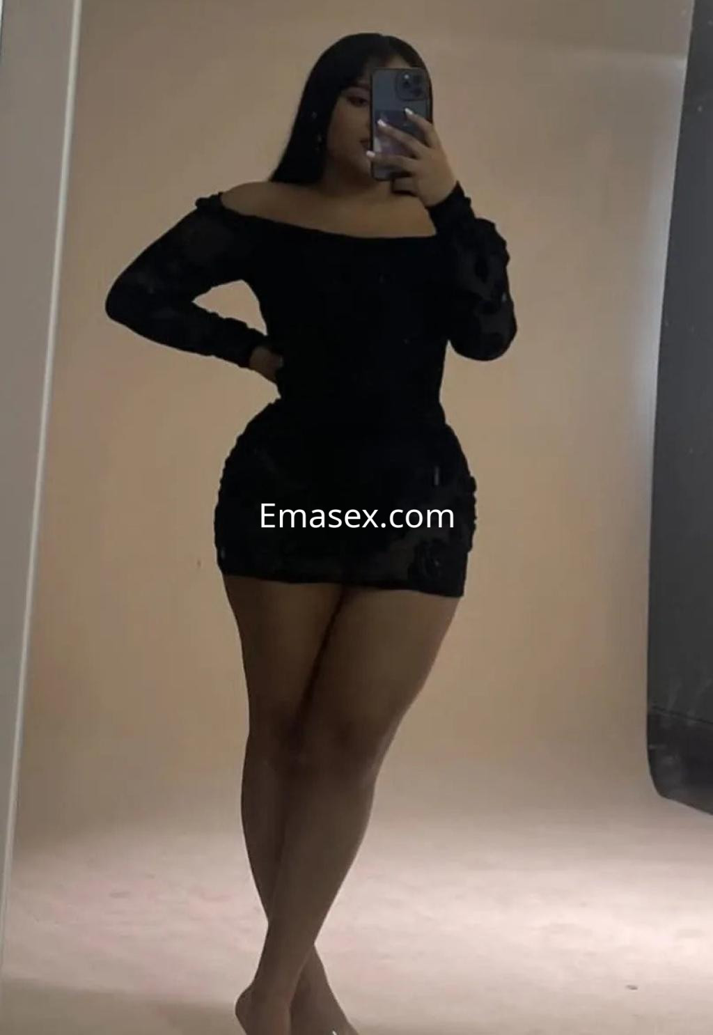 Escort en Cádiz, vistiendo un vestido negro de manga larga y ajustado, posa con confianza frente al espejo.