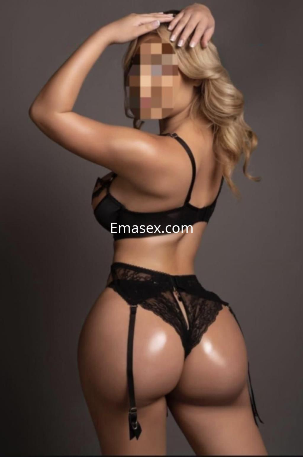 Escort en Cádiz posando de espaldas, con lencería negra y cabello rubio ondulado, en un fondo neutro.