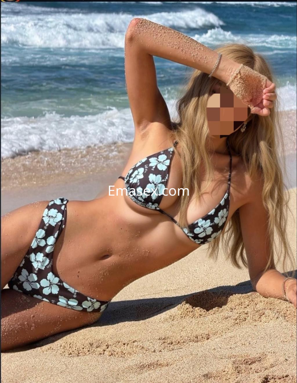 La escort está sentada en la playa, con un bikini de flores sobre fondo oscuro y el cabello suelto al viento.