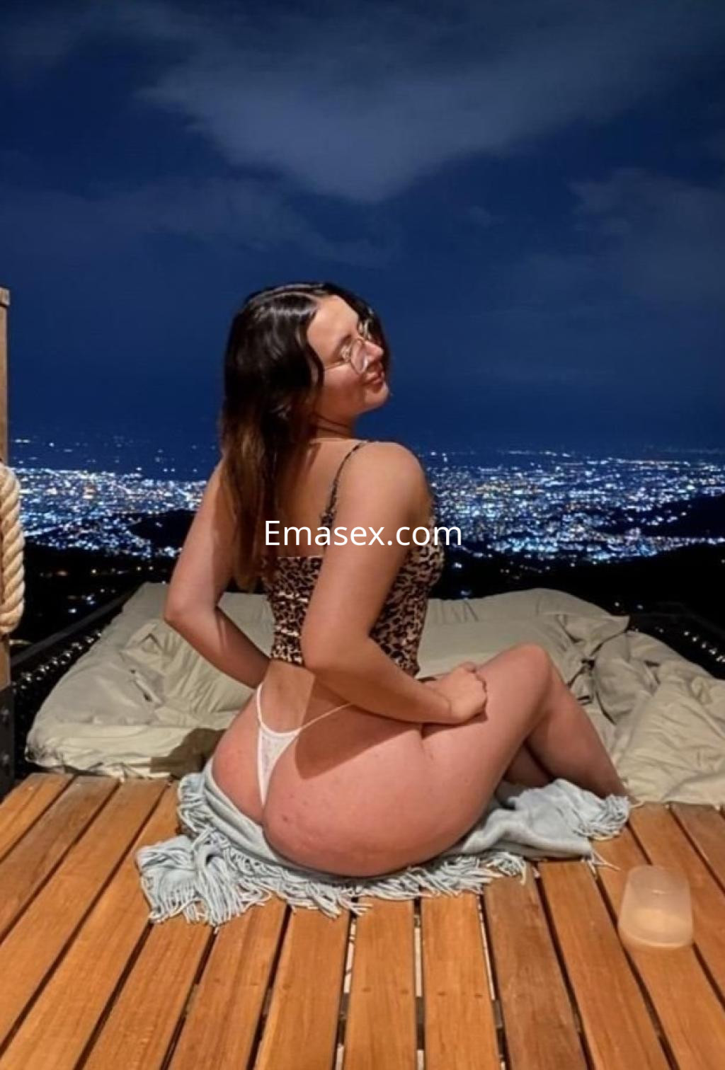El escort está sentada en una terraza de madera con vistas nocturnas de Málaga, vistiendo un bañador de estampado animal.